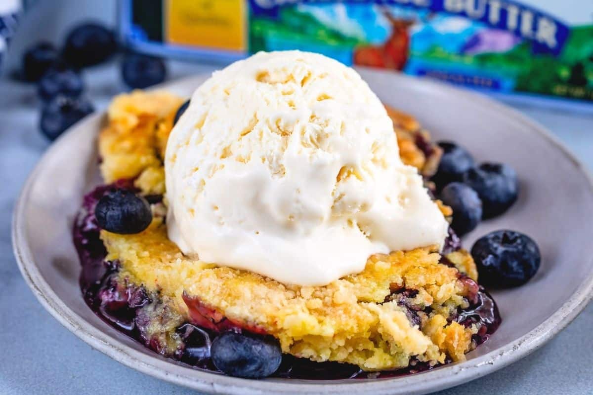 A scoop of vanilla ice cream sits on top of a golden blueberry dump cake cobbler, garnished with fresh blueberries and served on a gray plate.