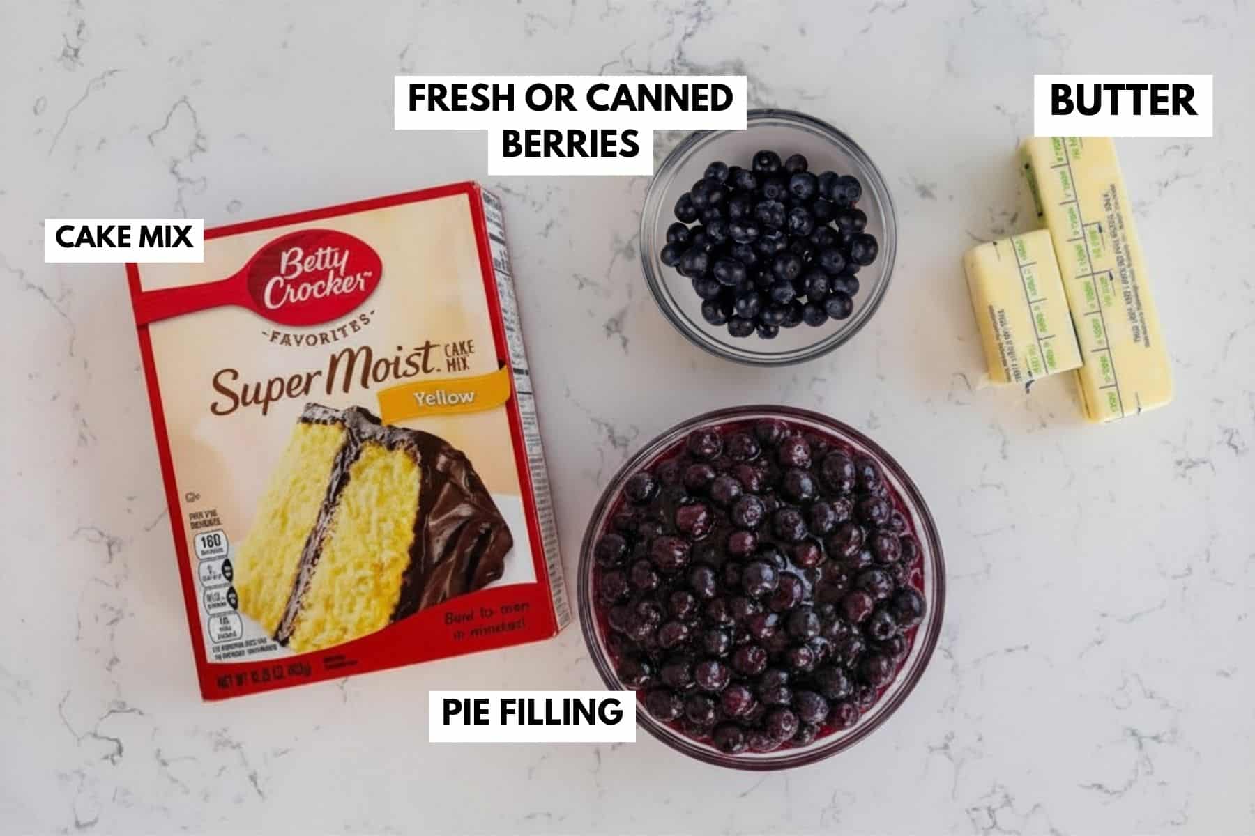 A box of yellow cake mix, a bowl of blueberries labeled fresh or canned berries, two sticks of butter, and a bowl labeled pie filling are arranged on a white surface—everything you need to make a delicious blueberry dump cake.
