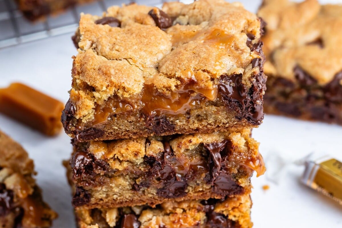 A close-up of three stacked chocolate chip cookie bars filled with gooey caramel and melted chocolate chips, with a soft and crumbly texture. Unwrapped caramel candies are visible in the background.