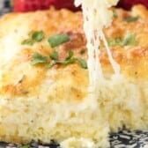 A close-up of a cheesy, golden-brown egg casserole garnished with chopped herbs, with a fork lifting a gooey, melted cheese portion. Strawberries are blurred in the background.