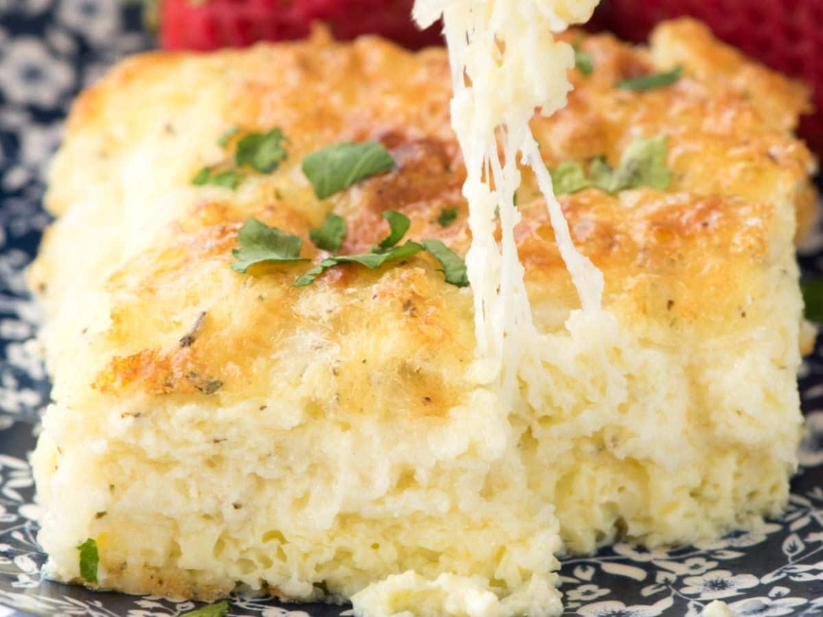 A close-up of a cheesy, golden-brown egg casserole garnished with chopped herbs, with a fork lifting a gooey, melted cheese portion. Strawberries are blurred in the background.