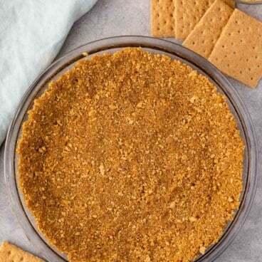 A pie dish with a golden-brown graham cracker crust sits on a gray surface. Beside it are several whole graham crackers and a small bowl of crushed crumbs. A light blue cloth is draped nearby.