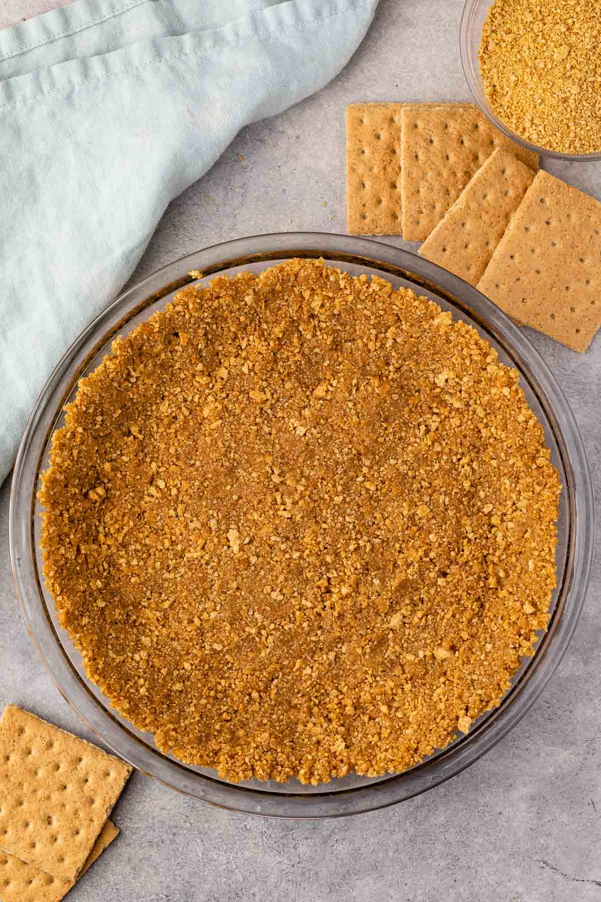 A pie dish with a golden-brown graham cracker crust sits on a gray surface. Beside it are several whole graham crackers and a small bowl of crushed crumbs. A light blue cloth is draped nearby.