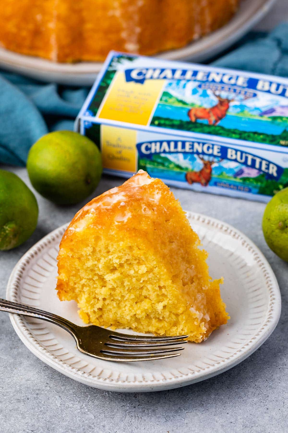 slicer of cake on white plate with fork and limes behind and butter box.