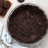 A glass pie dish filled with a rich Oreo crust recipe sits on a gray surface next to whole sandwich cookies on a wooden board, a striped kitchen towel, and a metal pie server.