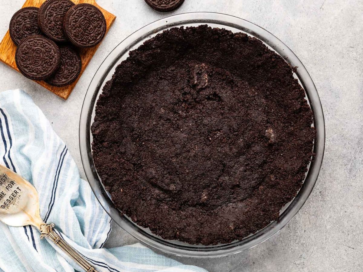 A glass pie dish filled with a rich Oreo crust recipe sits on a gray surface next to whole sandwich cookies on a wooden board, a striped kitchen towel, and a metal pie server.