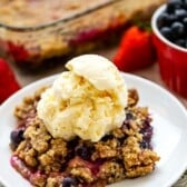 berry crisp on a white plate mixed with various berries with ice cream dolloped on top.