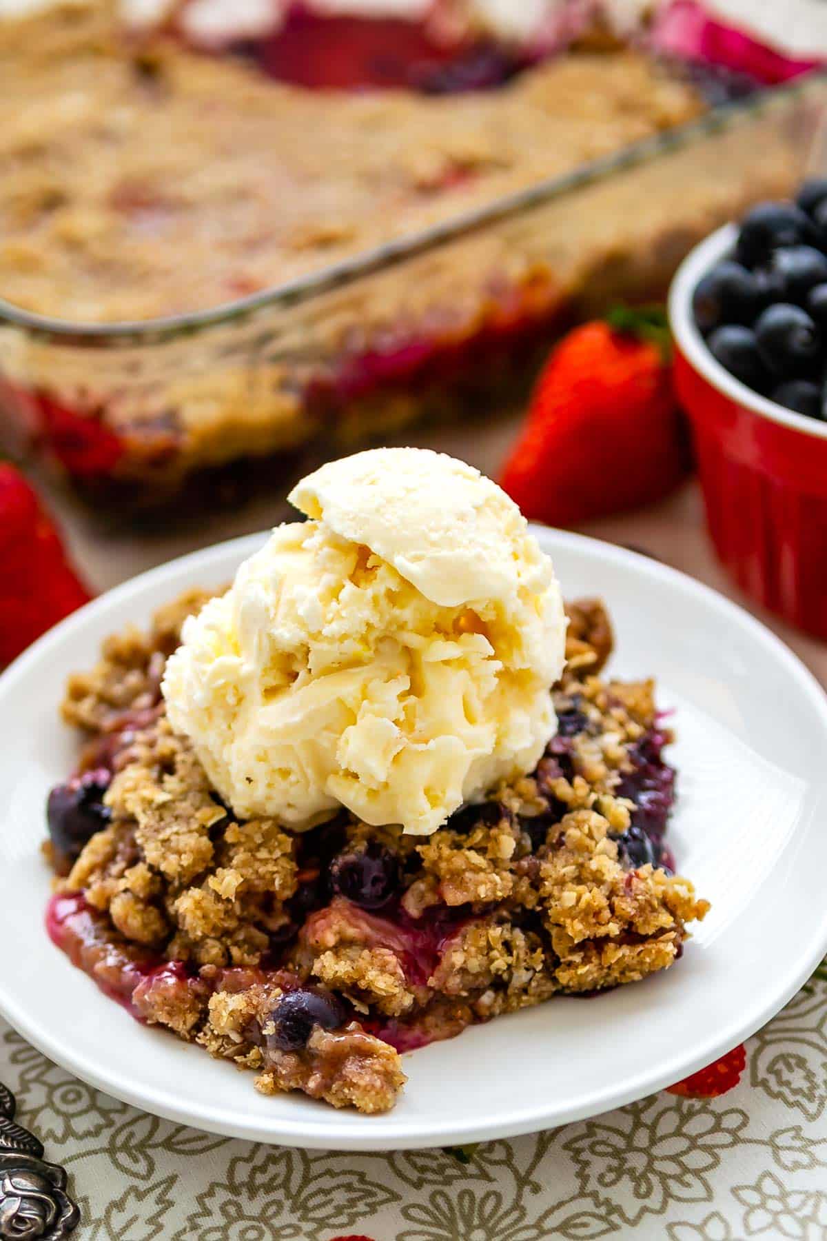 berry crisp on a white plate mixed with various berries with ice cream dolloped on top.