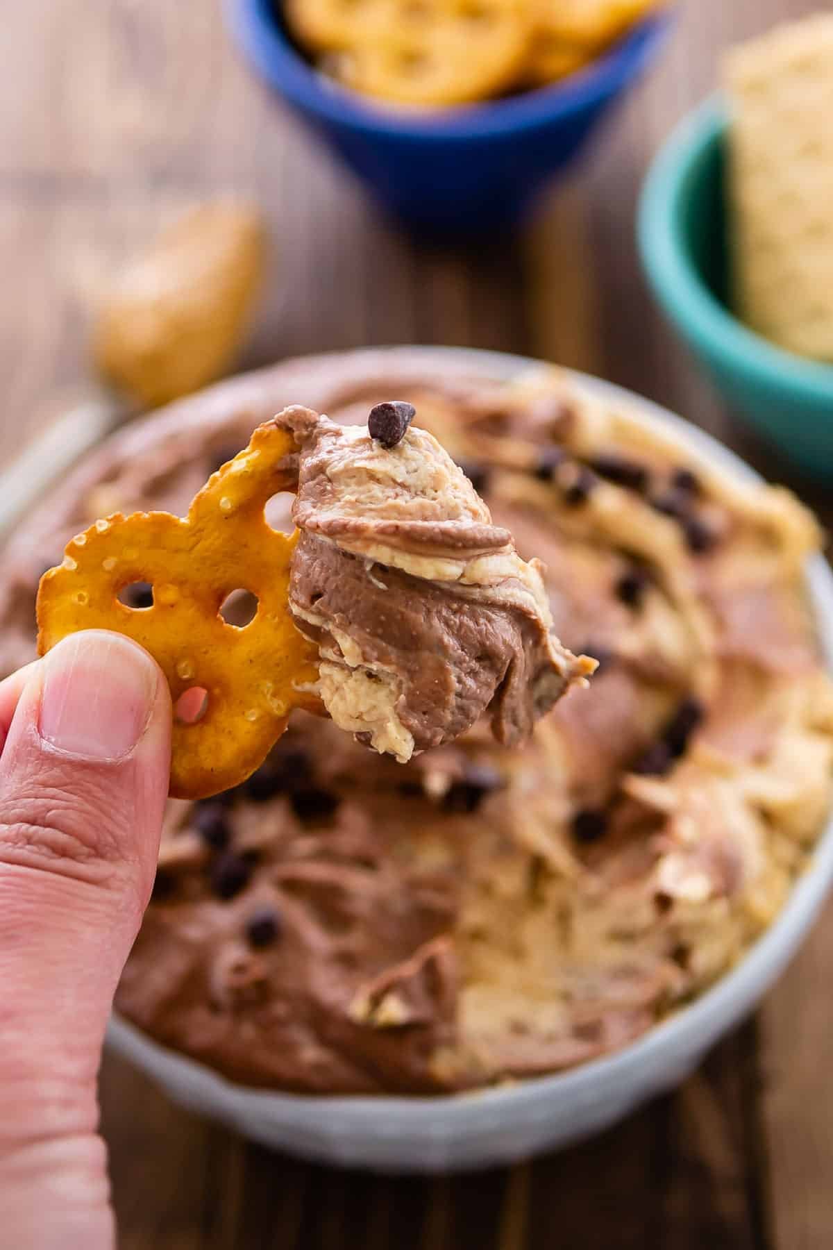 dip in a white bowl with mini chocolate chips sprinkles on top with a hand holding a pretzel over it.