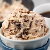 A close-up of a bowl of creamy coffee ice cream with chocolate chunks, placed in a white bowl with a blue base. A cup of coffee and an ice cream container are blurred in the background.