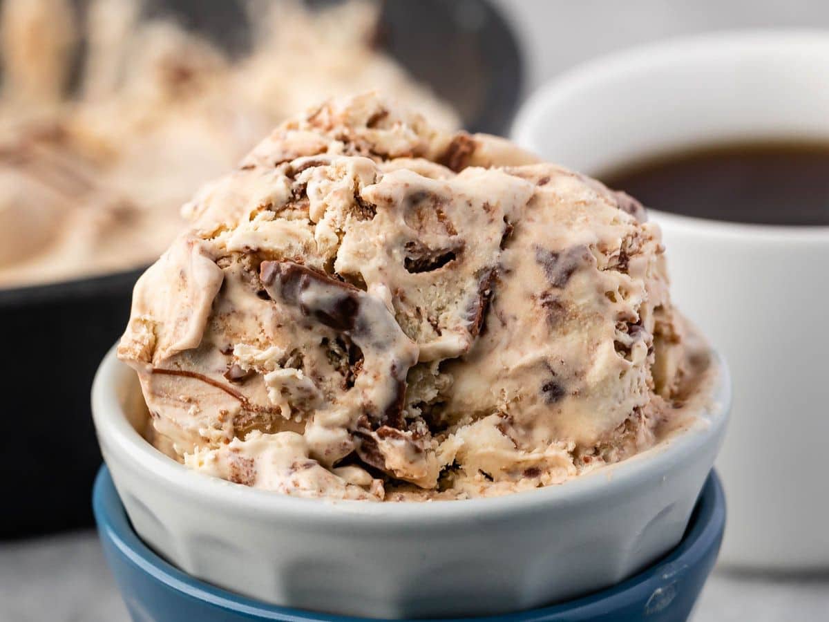 Gros plan d'un bol de glace au café crémeuse avec des pépites de chocolat, placé dans un bol blanc à fond bleu. Une tasse de café et un pot de glace sont flous en arrière-plan.
