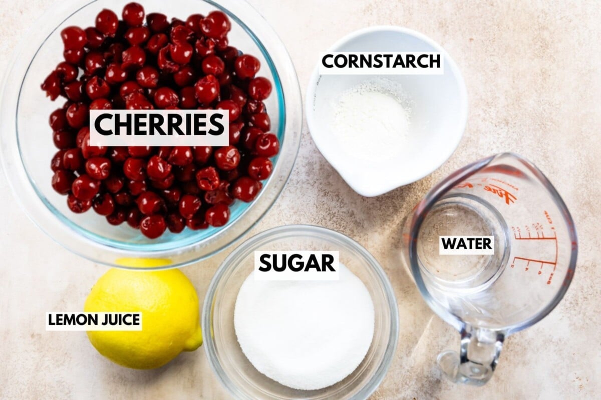A top view of bowls and containers with cherries, cornstarch, sugar, water, and a lemon labeled as lemon juice—essential ingredients for a classic cherry pie filling recipe—displayed on a light surface.
