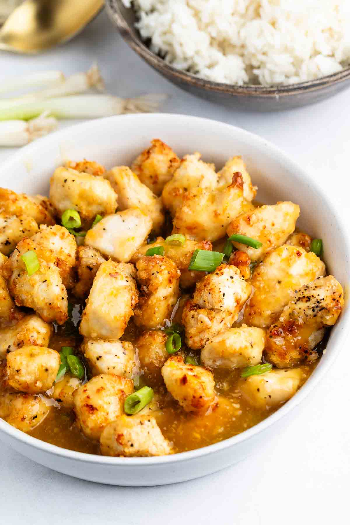 A bowl of golden, crispy Baked Orange Chicken pieces in a glossy sauce, garnished with chopped green onions, with a bowl of white rice and some green onions in the background.