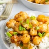 A plate of white rice topped with crispy, golden Baked Orange Chicken pieces coated in a glossy orange sauce and garnished with chopped green onions. A bowl of more chicken is blurred in the background.