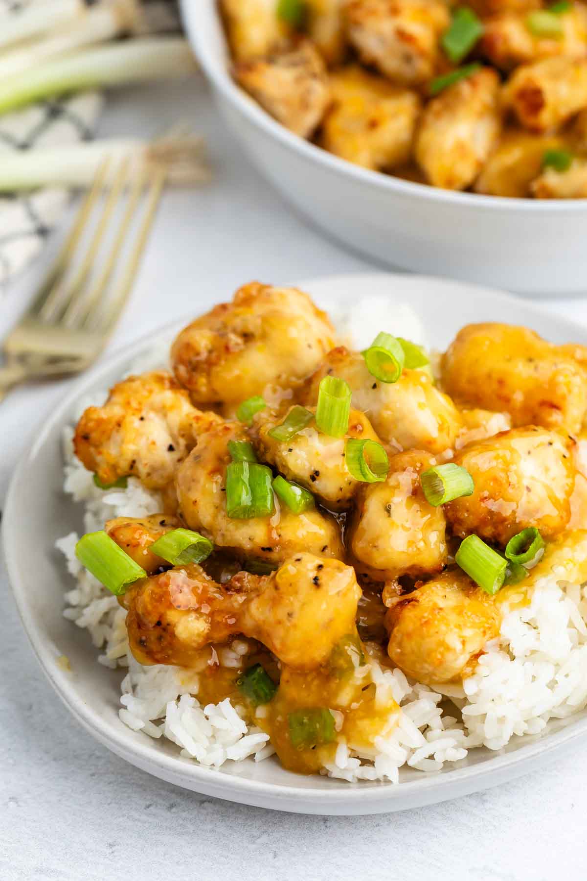 A plate of white rice topped with crispy, golden Baked Orange Chicken pieces coated in a glossy orange sauce and garnished with chopped green onions. A bowl of more chicken is blurred in the background.