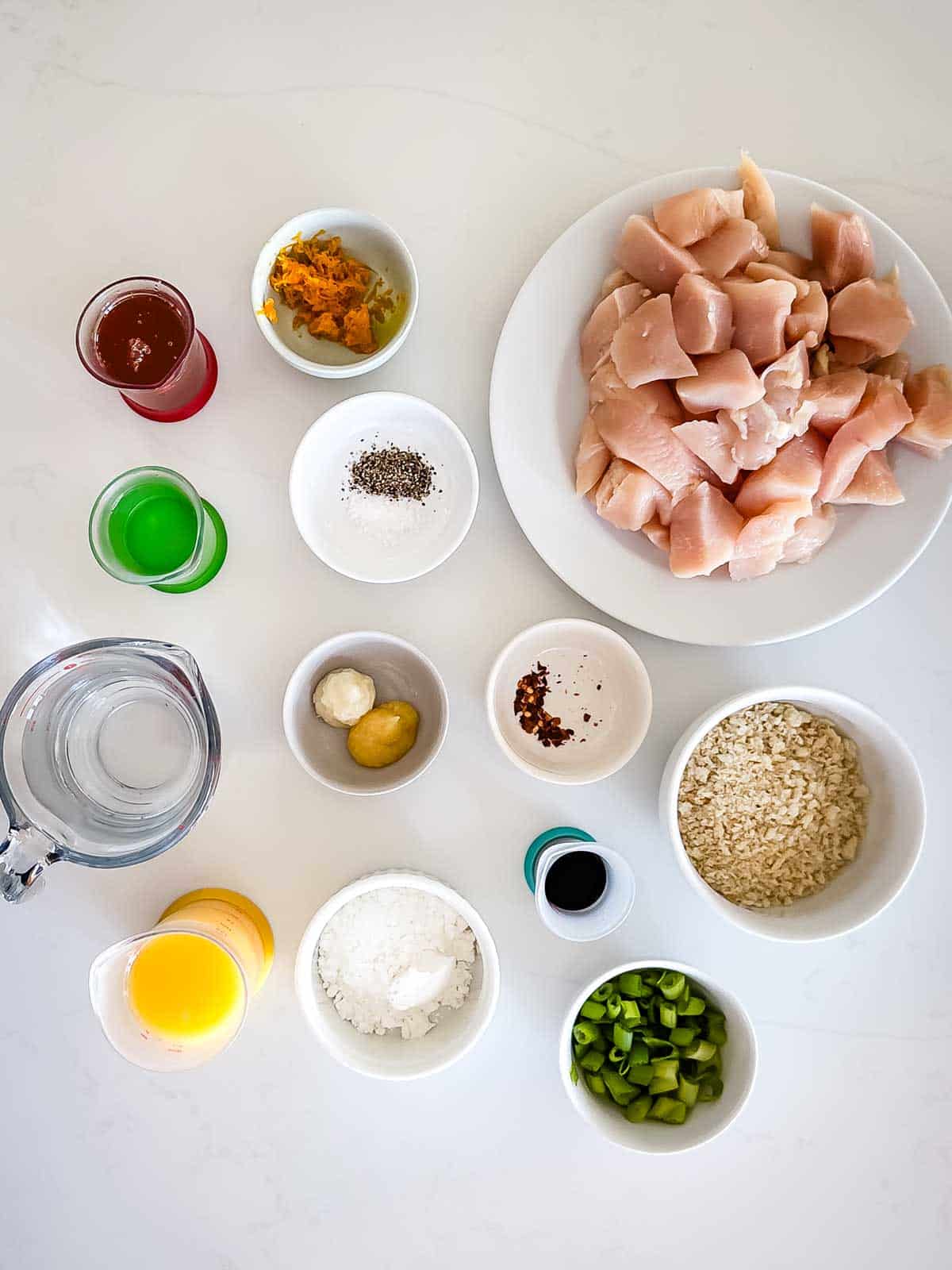 Overhead view of neatly arranged ingredients for Baked Orange Chicken on a white surface, including diced raw chicken, chopped green onions, orange juice, rice, flour, water, sauces, seasonings, and small bowls of garlic and ginger.