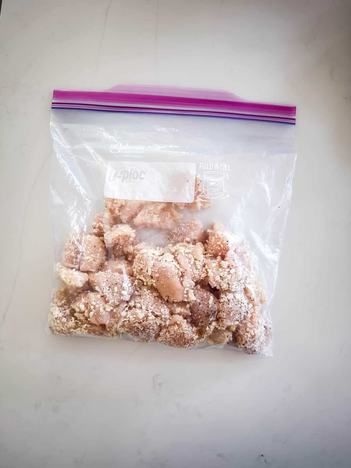 A clear Ziploc bag containing pieces of breaded, uncooked chicken for Baked Orange Chicken sits on a white countertop.