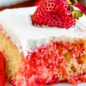 A slice of moist Jello Poke Cake topped with white whipped cream and a whole fresh strawberry, with strawberries on the side and red strawberry syrup seeping into the yellow cake.