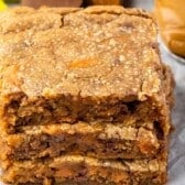 A close-up of three thick, golden-brown Peanut Butter Blondies stacked on parchment paper, with a spoonful of peanut butter and colorful candy pieces visible in the background.