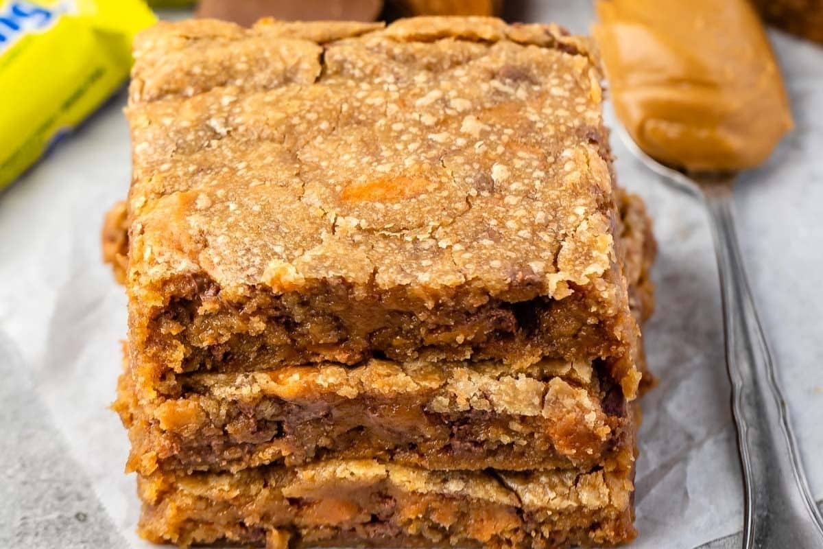 A close-up of three thick, golden-brown Peanut Butter Blondies stacked on parchment paper, with a spoonful of peanut butter and colorful candy pieces visible in the background.