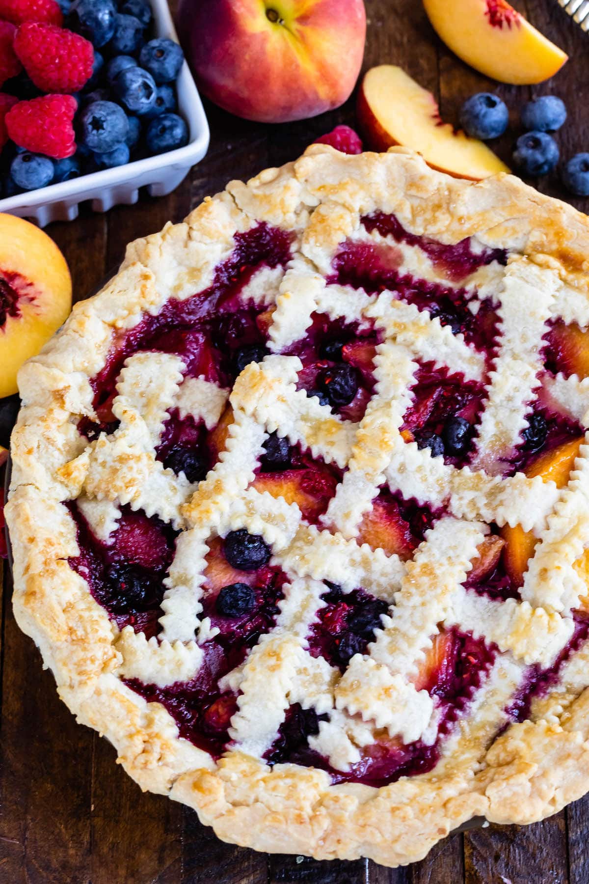 berry pie with decorative pie crust on top surrounded by slice fruit.