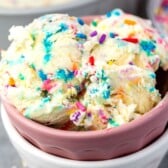 Scoops of cake batter ice cream with colorful rainbow sprinkles are served in a pink bowl. A blurred bowl of sprinkles and another dish appear in the background.