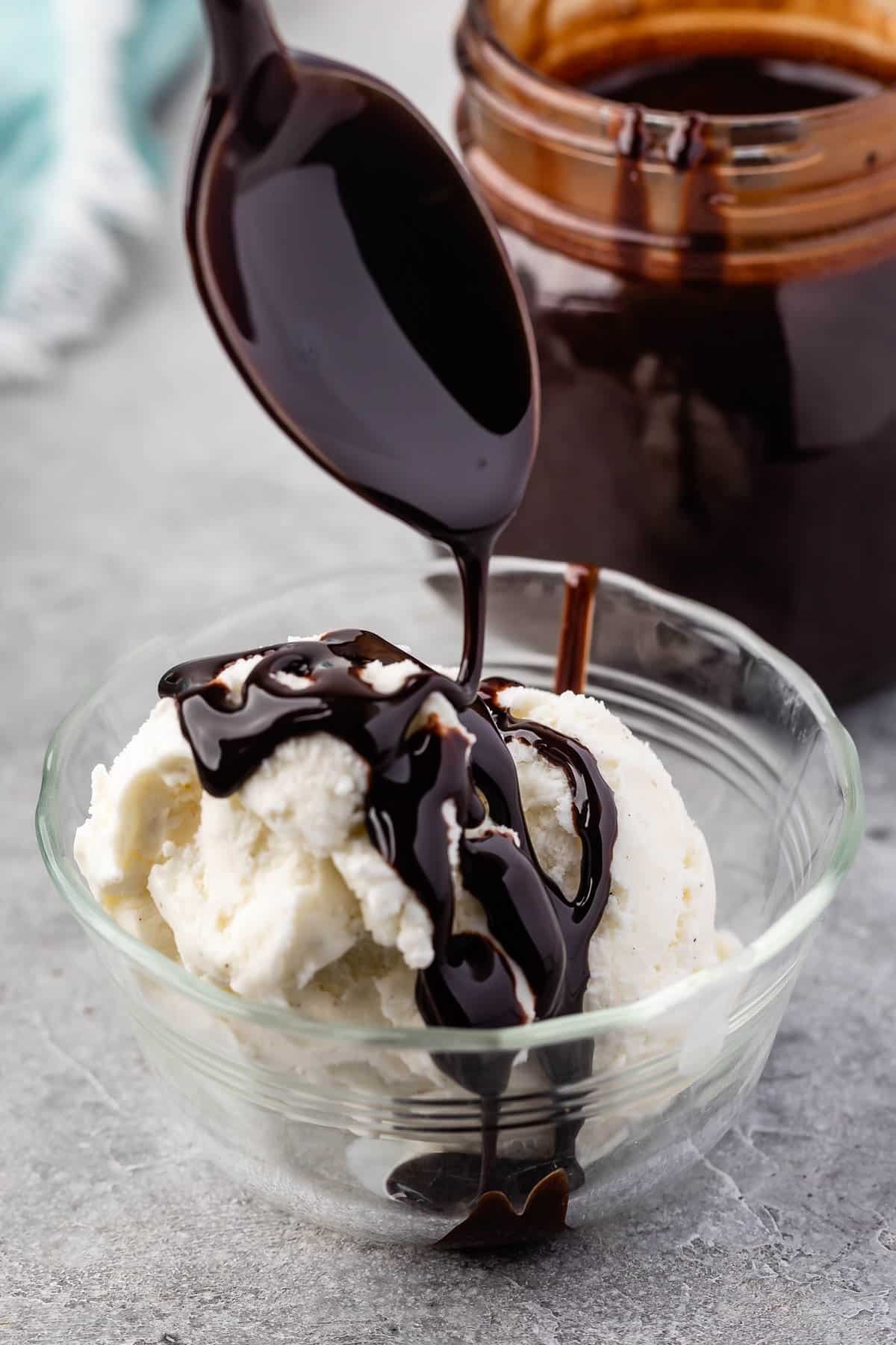 a spoon dripping chocolate sauce over vanilla ice cream in a clear bowl.