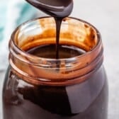 A spoon drizzles thick homemade chocolate sauce into a glass jar filled with the rich, glossy sauce. The jar&rsquo;s rim is coated with some chocolate, and a blue cloth is blurred in the background.