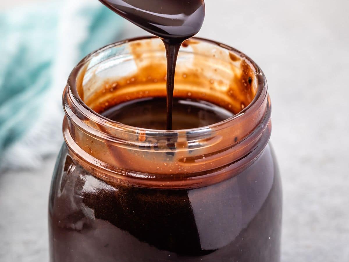 A spoon drizzles thick homemade chocolate sauce into a glass jar filled with the rich, glossy sauce. The jar’s rim is coated with some chocolate, and a blue cloth is blurred in the background.