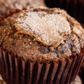 A close-up of a Chocolate Zucchini Muffin in a brown paper wrapper, topped with a sprinkle of sugar. The muffin appears moist and textured, with cracks on the surface. Other chocolate zucchini muffins are blurred in the background.