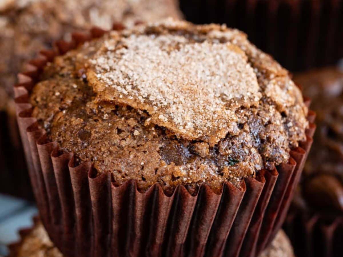 A close-up of a Chocolate Zucchini Muffin in a brown paper wrapper, topped with a sprinkle of sugar. The muffin appears moist and textured, with cracks on the surface. Other chocolate zucchini muffins are blurred in the background.