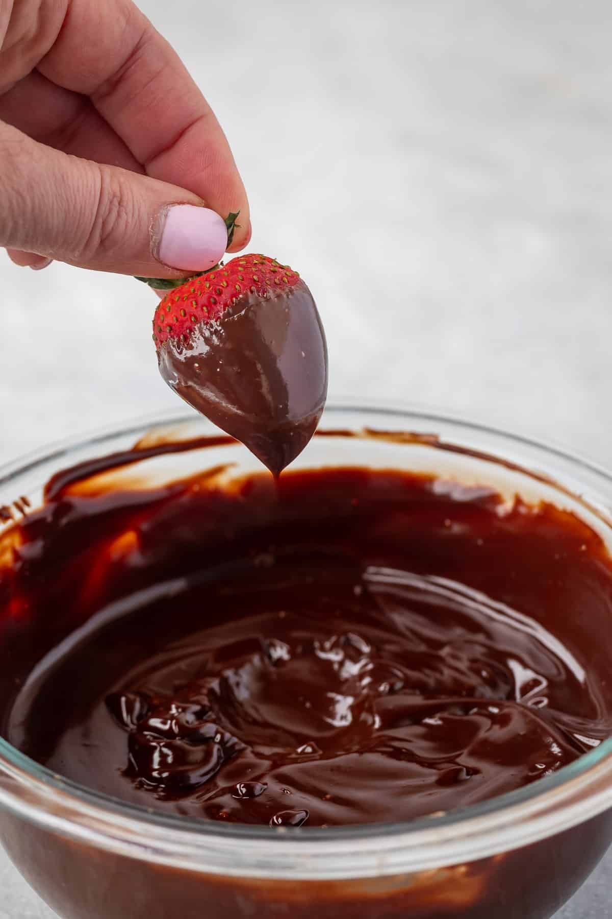 hand dipping strawberry into ganache in a clear glass bowl.