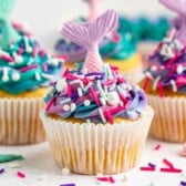 A vanilla mermaid cupcake with blue, pink, and purple swirled frosting, topped with colorful sprinkles and a pink mermaid tail decoration in a white paper liner. Other mermaid cupcakes are similarly decorated in the background.