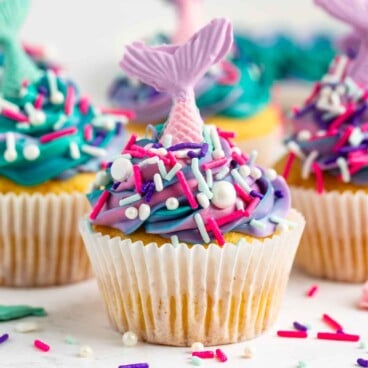 A vanilla mermaid cupcake with blue, pink, and purple swirled frosting, topped with colorful sprinkles and a pink mermaid tail decoration in a white paper liner. Other mermaid cupcakes are similarly decorated in the background.