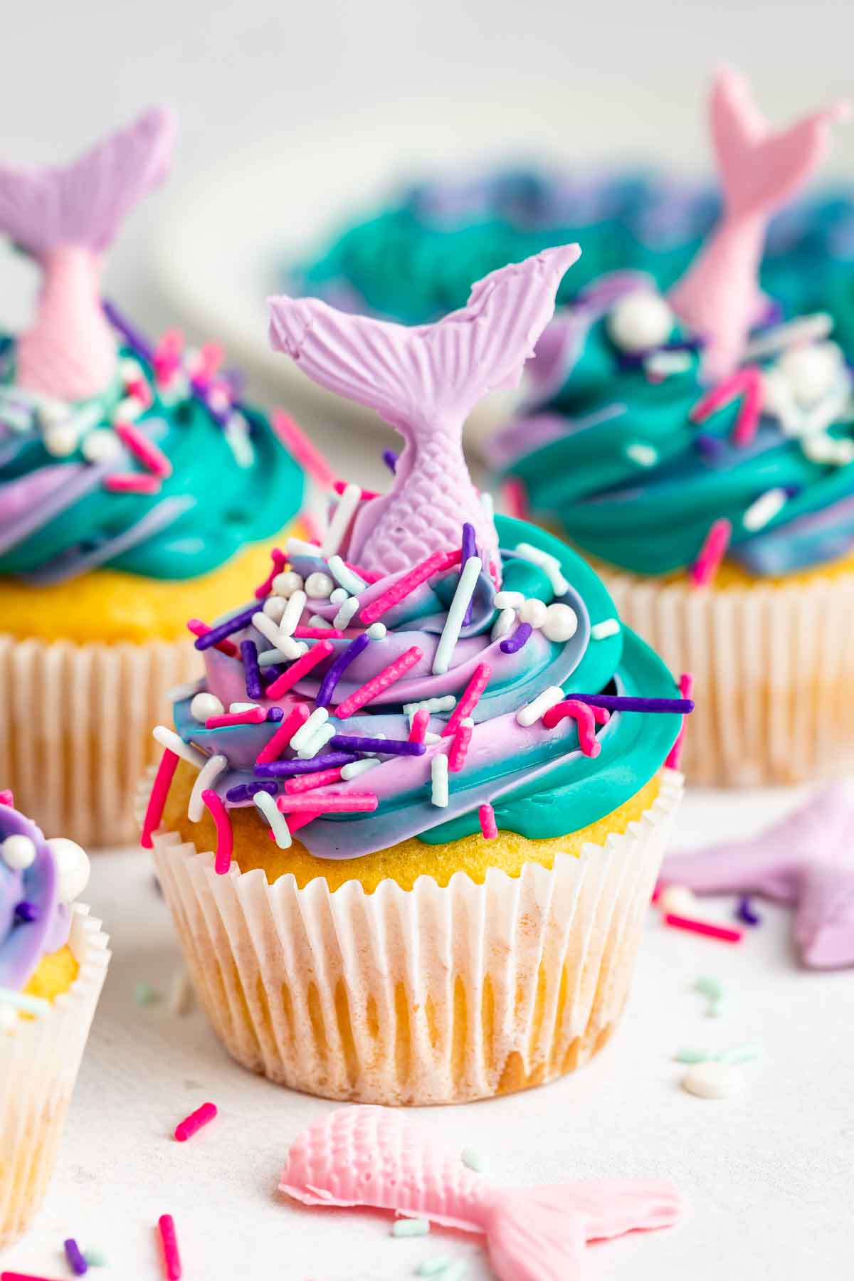 A mermaid cupcake decorated with swirled blue, purple, and teal frosting, colorful sprinkles, and a pink candy mermaid tail. Other mermaid cupcakes and candy tails add to the whimsical scene in the background.
