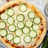 A freshly baked zucchini pizza topped with melted cheese and evenly spaced slices of zucchini, placed on parchment paper next to a small bowl of additional zucchini slices and a whole zucchini.