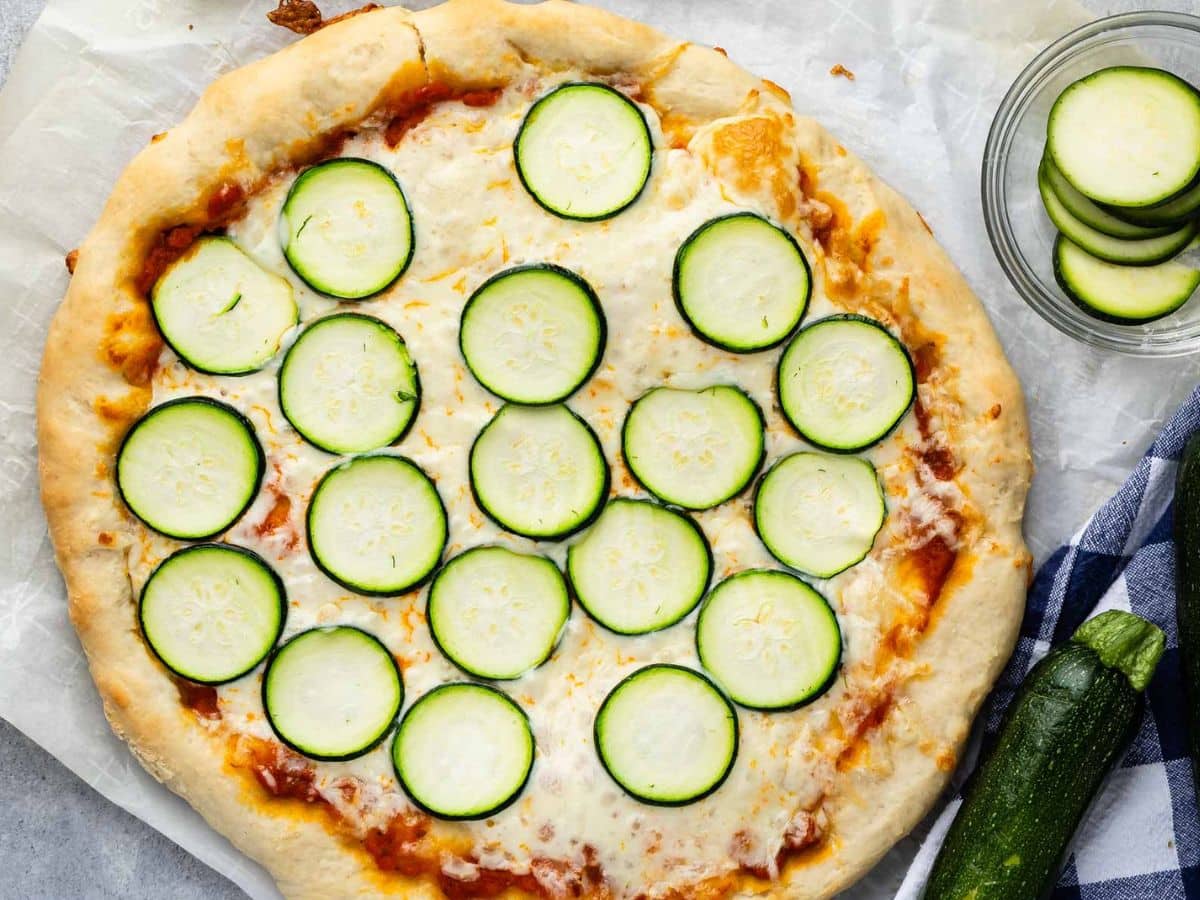 A freshly baked zucchini pizza topped with melted cheese and evenly spaced slices of zucchini, placed on parchment paper next to a small bowl of additional zucchini slices and a whole zucchini.