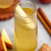 yellow drink in a tall clear glass with an apple slice in the glass.