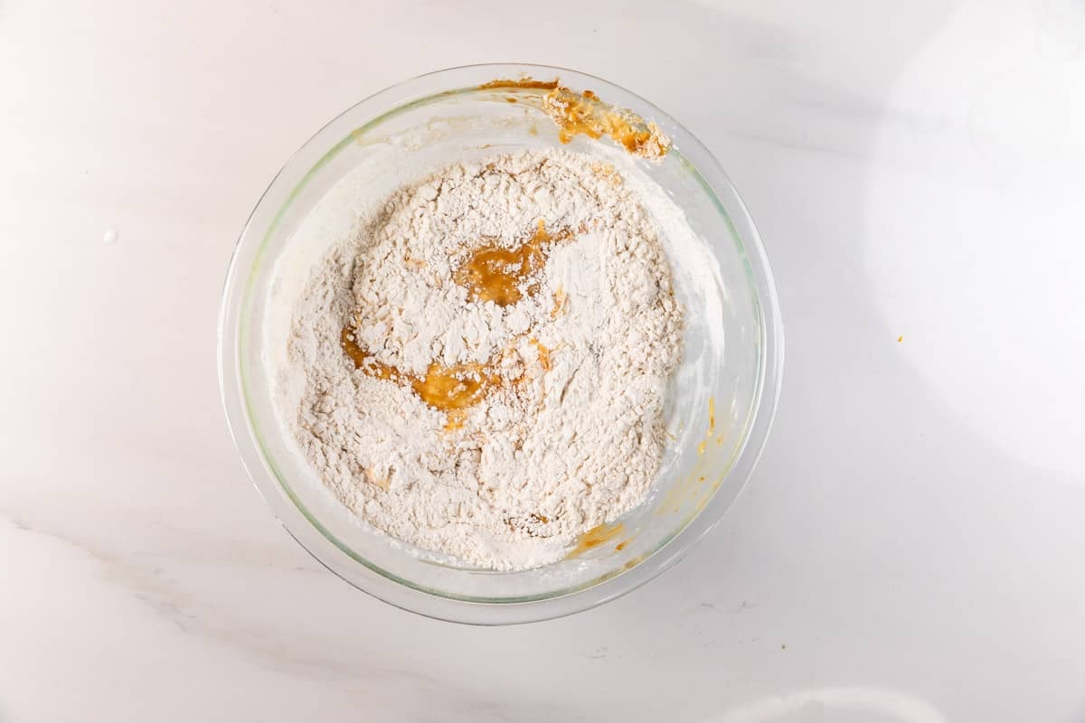 A glass mixing bowl on a white countertop contains flour and wet ingredients partially mixed together for Biscoff Banana Bread, showing patches of both dry and wet ingredients.