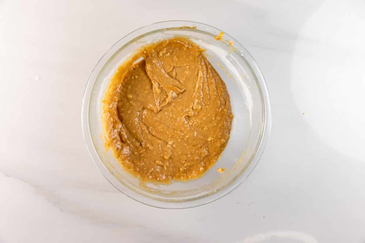 A glass bowl filled with thick, light brown Biscoff Banana Bread batter sits on a white marble surface. The mixture appears well-mixed and slightly textured, ready for baking a delicious twist on classic banana bread.