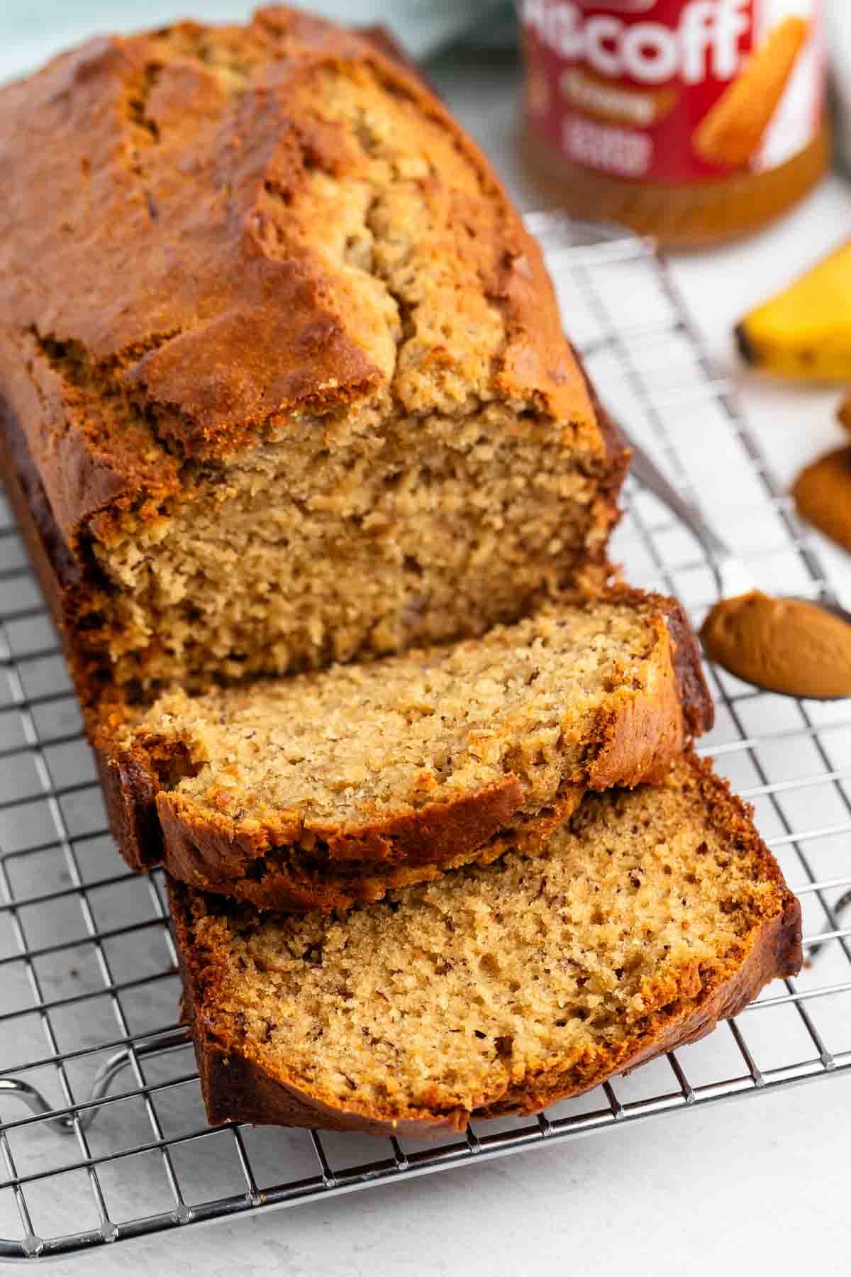 A loaf of Biscoff Banana Bread with two slices cut rests on a wire cooling rack. The bread looks moist with a golden-brown crust, and a jar of Biscoff spread is visible in the background.