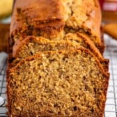 A loaf of homemade Biscoff Banana Bread with a golden-brown crust, partially sliced, sits on a wire cooling rack. The bread looks moist and dense, with visible banana pieces and hints of Biscoff spread inside.