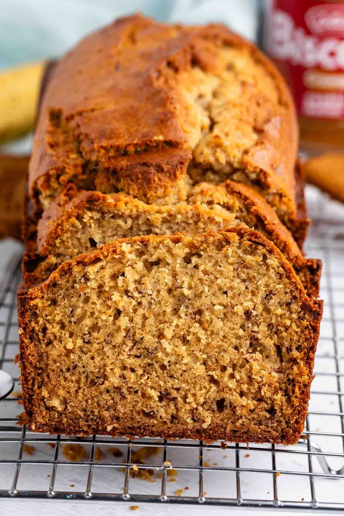 A loaf of homemade Biscoff Banana Bread with a golden-brown crust, partially sliced, sits on a wire cooling rack. The bread looks moist and dense, with visible banana pieces and hints of Biscoff spread inside.