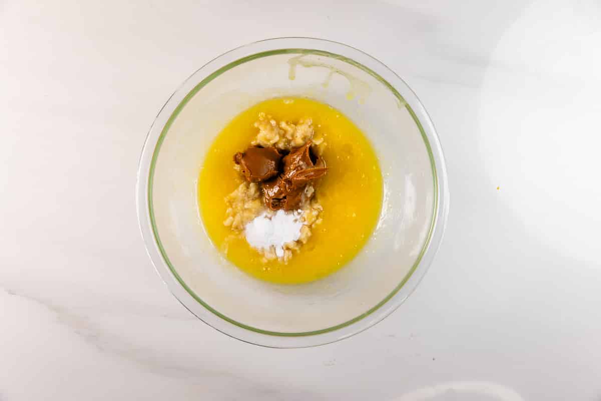A glass bowl on a white surface holds melted butter, mashed banana, dollops of peanut butter, Biscoff spread, and a small pile of baking soda—ingredients ready to be mixed for delicious Biscoff Banana Bread.