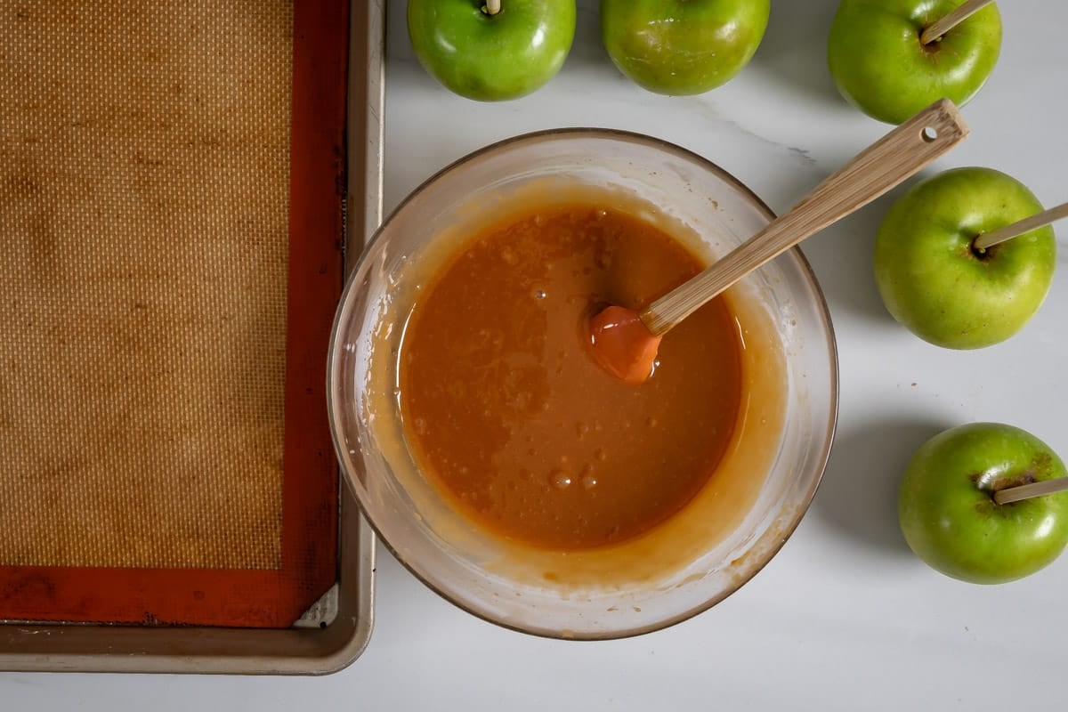 clear bowl of caramel with a spatula in it with apples around the bowl.