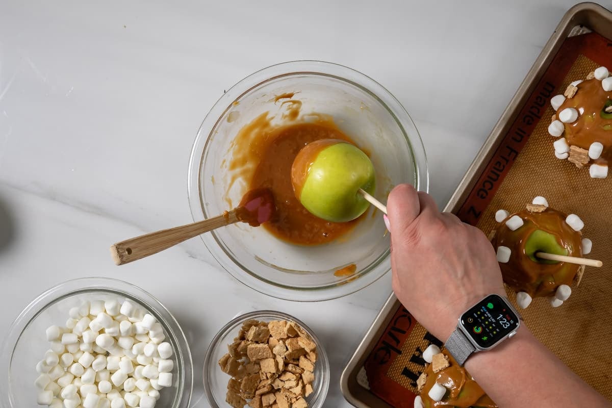 hand holding caramel apple of a skewer over a bowl of caramel.