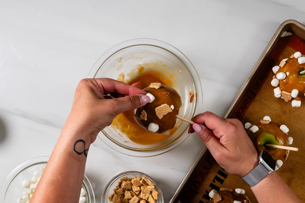 hand holding caramel apple of a skewer over a bowl of caramel.