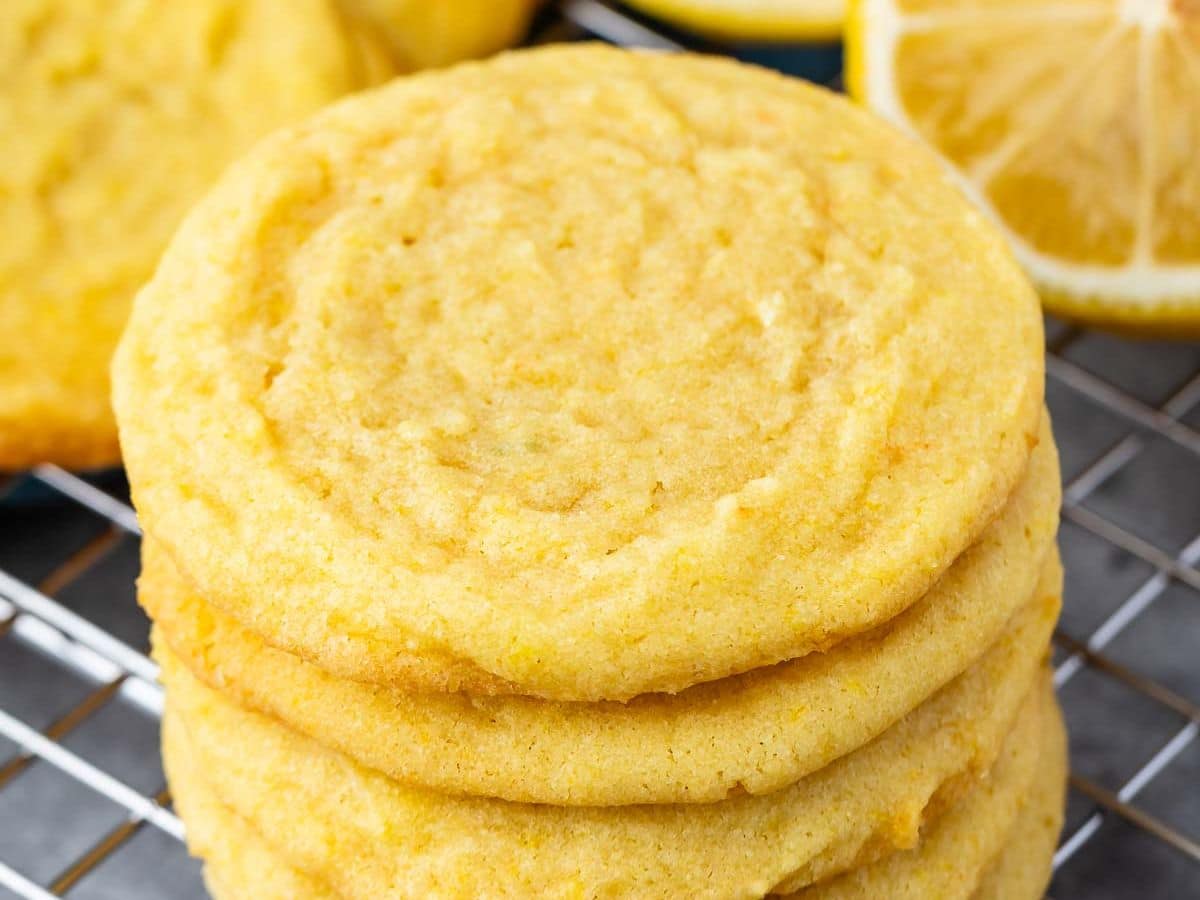A close-up of a stack of golden-yellow Lemon Pudding Cookies resting on a cooling rack, with a sliced lemon visible in the background.