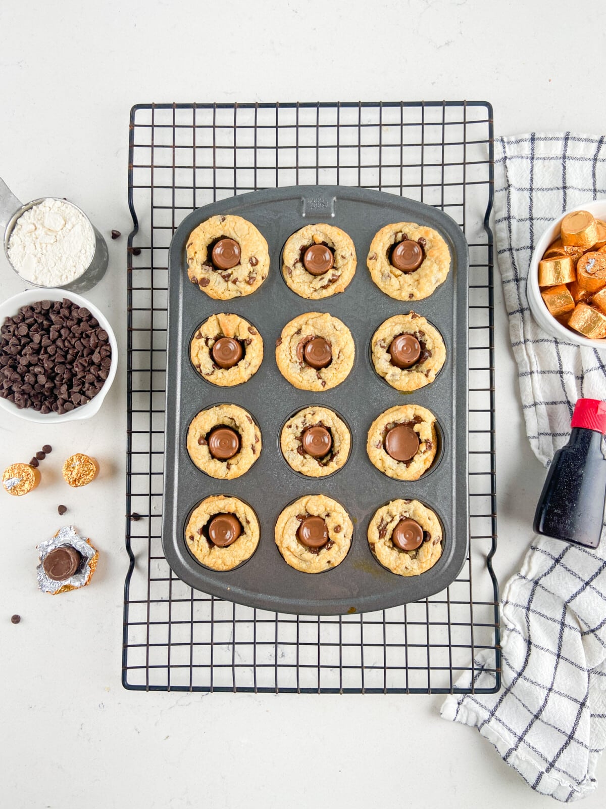 A muffin tin with 12 freshly baked rolo cookies sits on a cooling rack, each cookie cup holding a chocolate candy center. Nearby are bowls of chocolate chips, flour, caramel candies, and a bottle of vanilla extract.