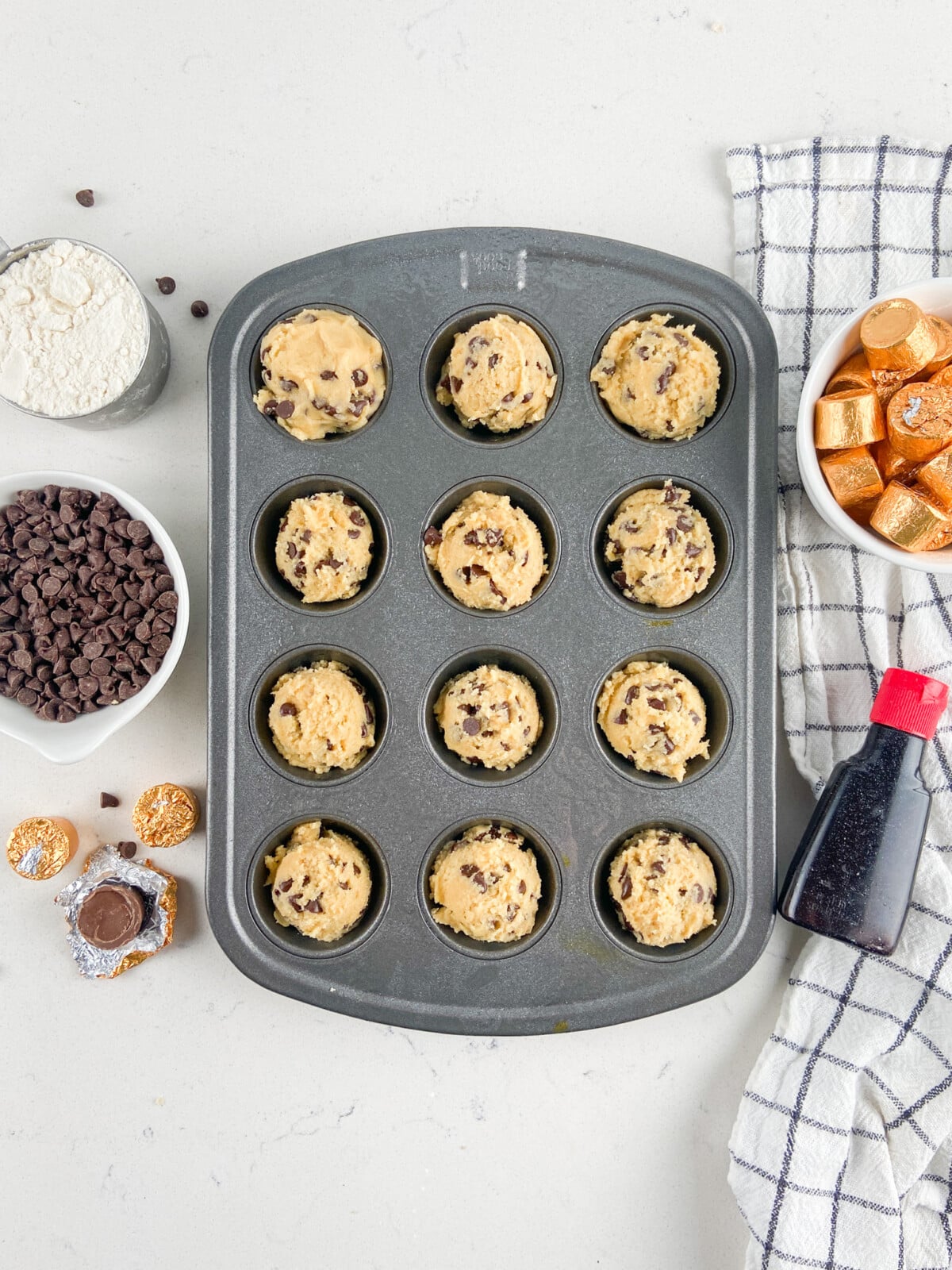 A muffin tin filled with chocolate chip cookie dough sits on a white surface, surrounded by bowls of chocolate chips, flour, wrapped caramel candies for rolo cookies, vanilla extract, and a striped kitchen towel.